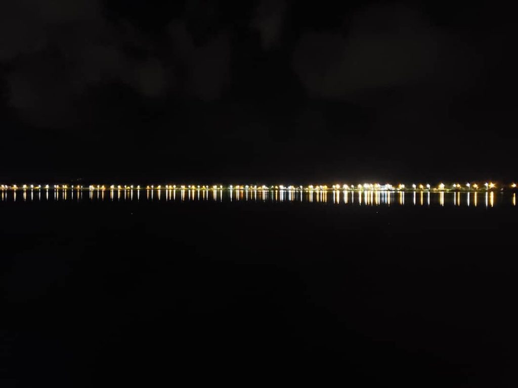 Na imagem, vemos luzes ao longe, refletindo em uma lagoa durante a noite